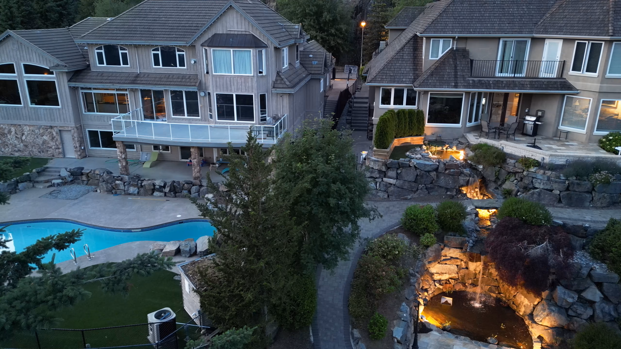 A lit stone waterfall, pool and terraced walls at dusk, from the air